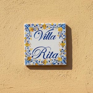 May include: Square ceramic tile with the words "Villa Rita" in elegant blue script. The tile is white with a decorative border of yellow flowers and blue accents. The tile is mounted on a light yellow wall.