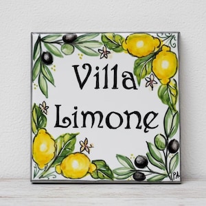 以下が含まれることがあります： 黒い筆記体で「Villa Limone」と書かれた正方形のセラミックタイル。タイルは、黄色のレモン、緑の葉、黒いオリーブ、小さな白い花で飾られています。デザインは緑と白のボーダーで縁取られています。