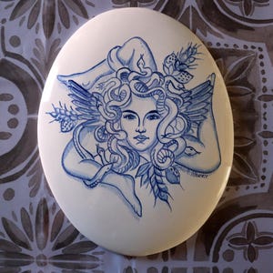 May include: Oval ceramic art piece featuring a blue and white illustration of a woman with snakes for hair, wings, and floral accents. The artwork is signed "F. Albanese".