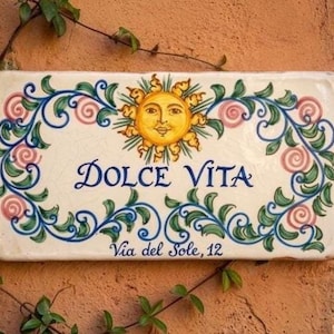 以下が含まれることがあります： 青い筆記体で「Dolce Vita」と書かれた長方形のセラミックサイン、笑顔の太陽、花のボーダー。サインには「Via del Sole, 12」の文字も含まれています。サインは、テクスチャ加工されたオレンジブラウンの壁に取り付けられています。