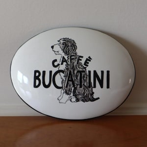 May include: Oval white sign with a black border featuring the words "CAFFE BUCCATINI" and a black and white illustration of a dog. The sign is likely for a cafe or restaurant.