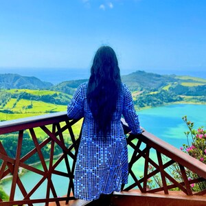 May include: A person stands on a wooden observation deck, overlooking a scenic landscape of green hills and a turquoise lake. The person wears a blue and white checkered dress. The sky is clear blue.