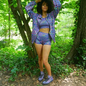 May include: A woman wearing a blue and pink geometric patterned jacket, matching shorts, and high-top sneakers. She is standing in a forest setting.