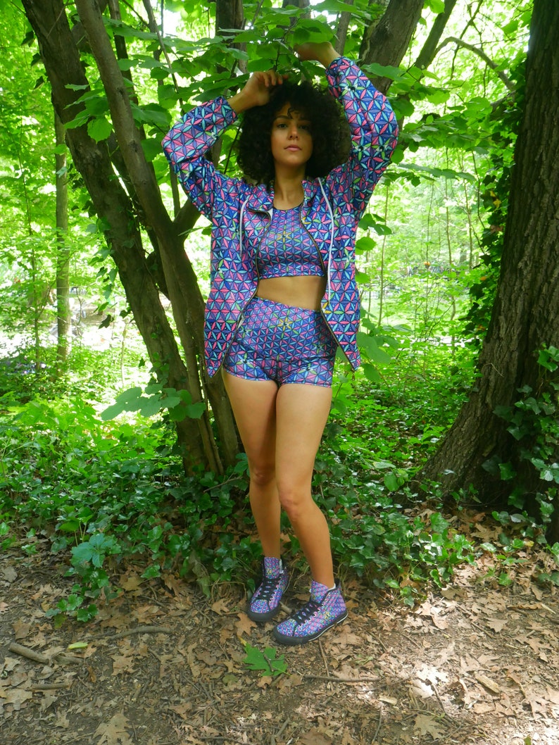 May include: A woman wearing a blue and pink geometric patterned jacket, matching shorts, and high-top sneakers. She is standing in a forest setting.