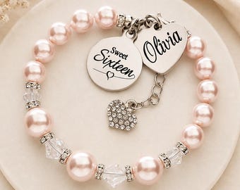 May include: A beaded bracelet featuring light pink pearls and clear crystal spacers. The bracelet includes a round silver charm engraved with Sweet Sixteen and a heart-shaped charm engraved with the name Olivia, plus a small crystal-encrusted heart charm.