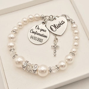 May include: White pearl bracelet with a silver charm that says "On your Confirmation 04/05/2023" and a silver heart charm that says "Olivia".