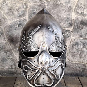 May include: A silver-colored helmet with an octopus design on the face and tentacles extending up the sides. The helmet has a pointed top and small rivets. The design is detailed and appears to be made of metal.