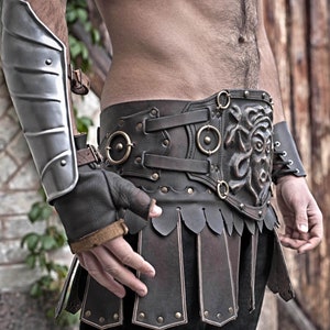 May include: A brown leather belt with a decorative metal buckle featuring a sculpted face. The belt has multiple straps and buckles, and is adorned with metal studs.