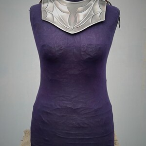 May include: A silver metal necklace with a decorative design. The necklace is on a purple mannequin.