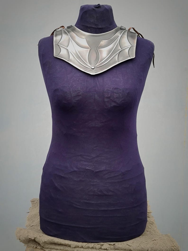 May include: A silver metal necklace with a decorative design. The necklace is on a purple mannequin.
