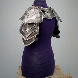 May include: Silver-toned metal shoulder armor with a spiderweb design. The armor is made of multiple plates and is secured with leather straps.