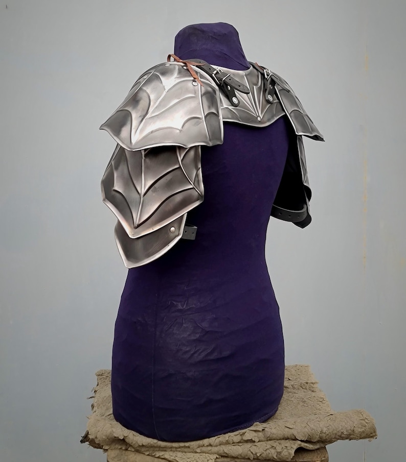 May include: Silver-toned metal shoulder armor with a spiderweb design. The armor is made of multiple plates and is secured with leather straps.
