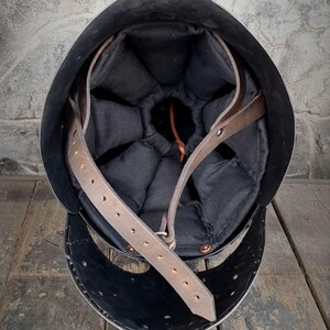 May include: A black metal helmet with a padded black interior and brown leather straps. The helmet is set against a rustic wooden surface and a textured gray wall.
