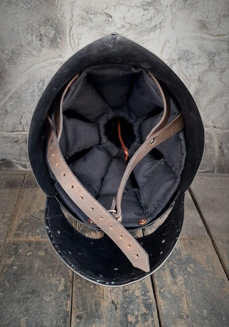 May include: A black metal helmet with a padded black interior and brown leather straps. The helmet is set against a rustic wooden surface and a textured gray wall.