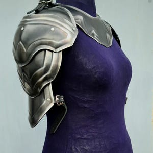 May include: A silver metal shoulder armour piece with a leather strap. The armour is worn on a mannequin with a purple shirt.