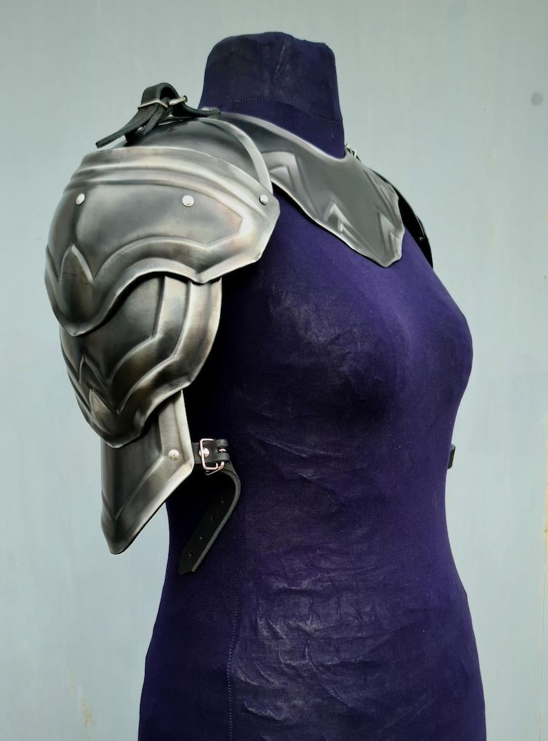 May include: A silver metal shoulder armour piece with a leather strap. The armour is worn on a mannequin with a purple shirt.