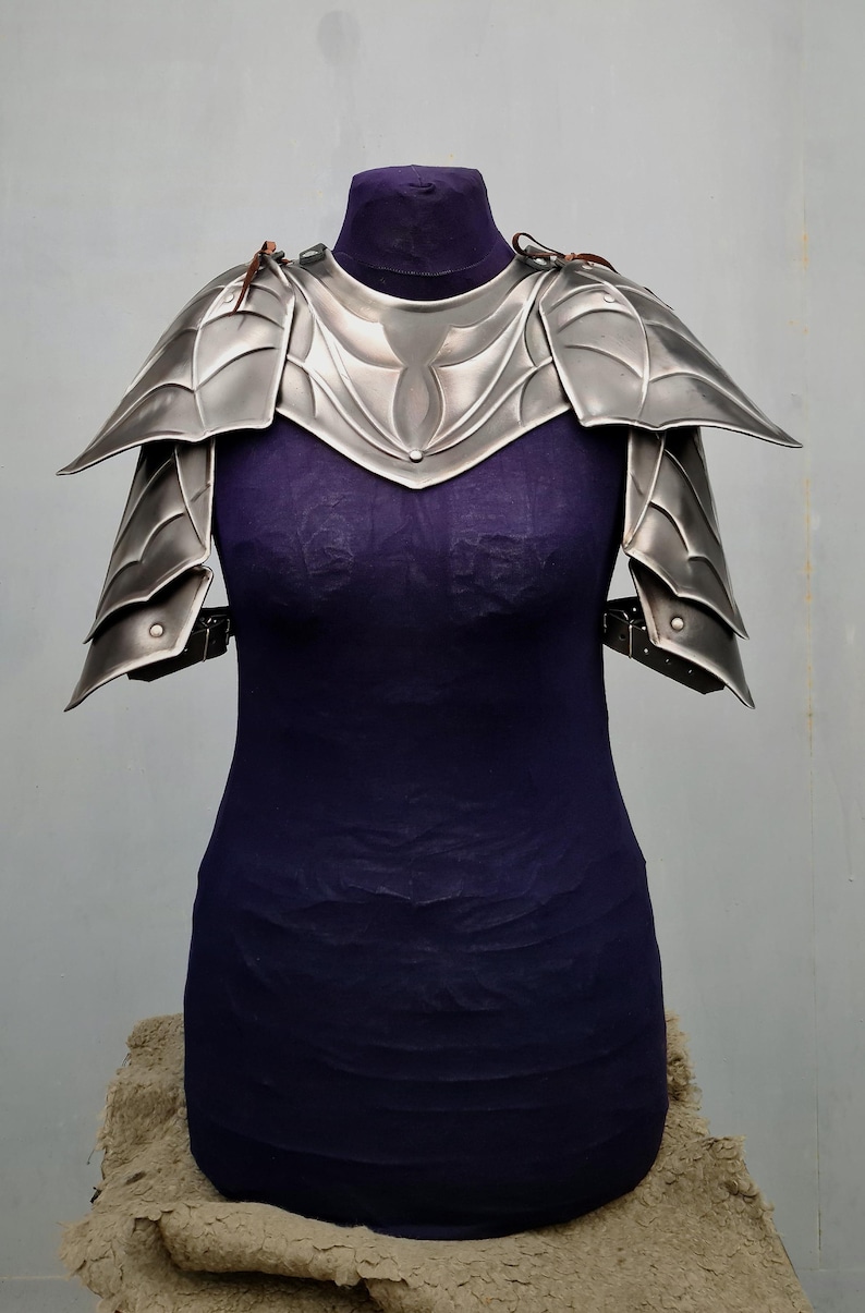 May include: Silver plated shoulder armor with a decorative design. The armor is made of metal and has a textured surface.