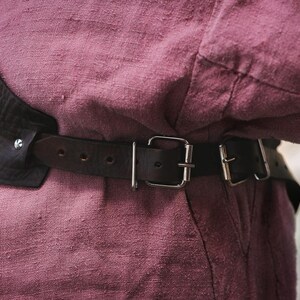 May include: A brown leather belt with silver buckles and a metal plate. The belt is worn over a pink shirt.