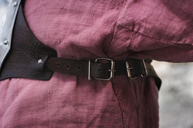 May include: A brown leather belt with silver buckles and a metal plate. The belt is worn over a pink shirt.