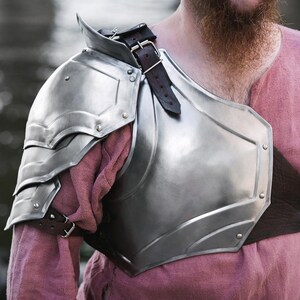 May include: A silver metal shoulder armor with a leather strap. The armor is worn over a pink shirt.