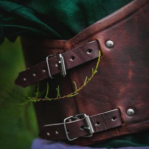 May include: A brown leather corset with silver buckles and rivets. The corset is worn over a green shirt and a purple skirt.