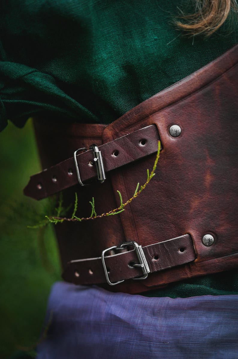 May include: A brown leather corset with silver buckles and rivets. The corset is worn over a green shirt and a purple skirt.