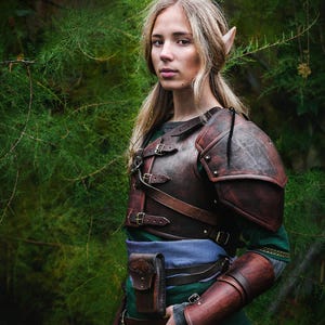 May include: A woman with pointed ears wearing a brown leather armor with straps and buckles. She is standing in a forest setting with green foliage.