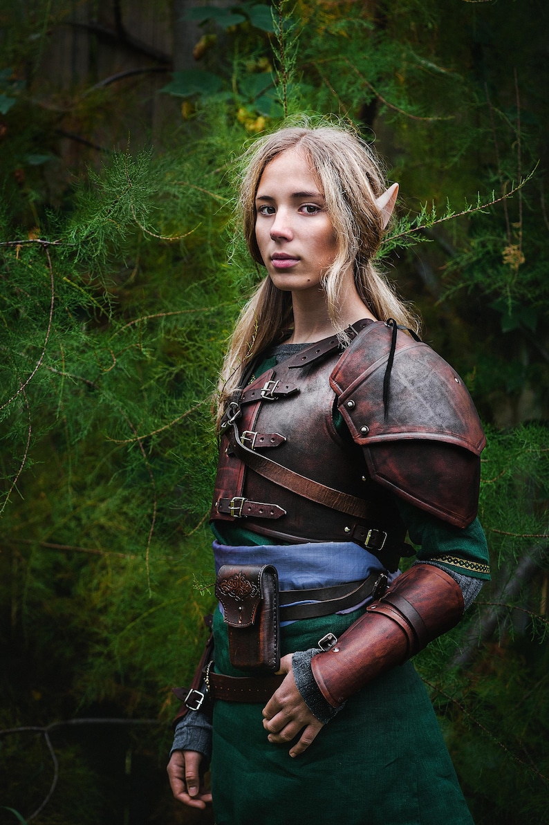 May include: A woman with pointed ears wearing a brown leather armor with straps and buckles. She is standing in a forest setting with green foliage.