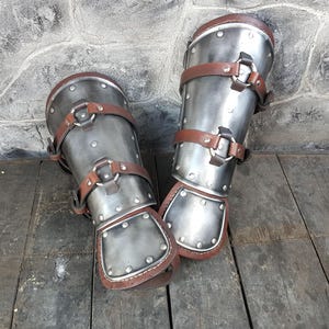 Op de afbeelding: Een paar zilveren metalen scheenbeschermers met bruine leren banden en gespen. De scheenbeschermers liggen op een houten oppervlak.