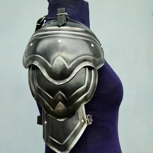 May include: A silver metal shoulder armour with a leather strap. The armour is made of multiple plates and has a decorative design.