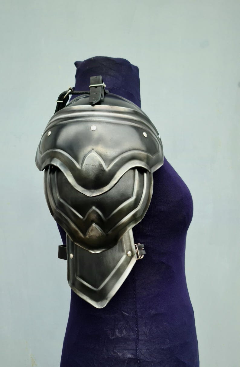 May include: A silver metal shoulder armour with a leather strap. The armour is made of multiple plates and has a decorative design.