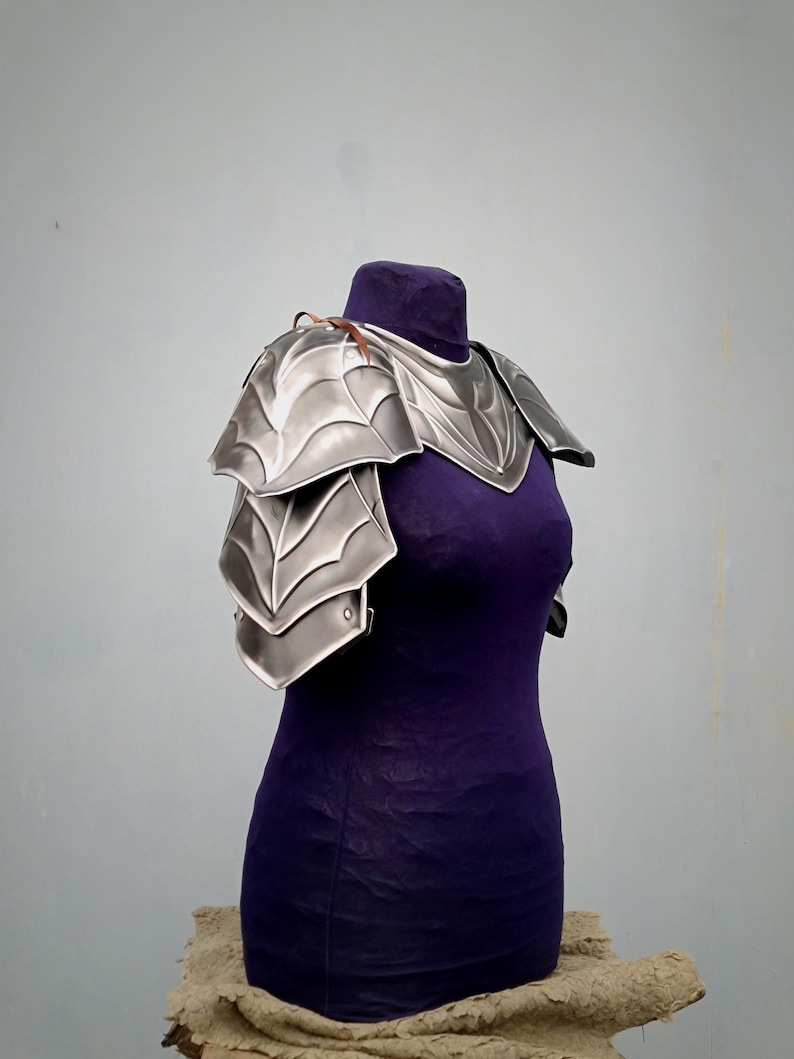 May include: Silver plated shoulder armor with a decorative design. The armor is displayed on a purple mannequin.