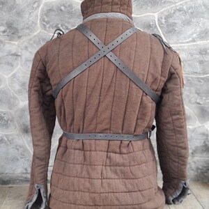 May include: A brown quilted jacket with a black leather strap across the back and a black leather belt around the waist.