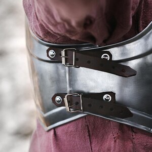 May include: Close-up of a silver metal chest plate with two brown leather straps and silver buckles. The chest plate is worn over a burgundy shirt.