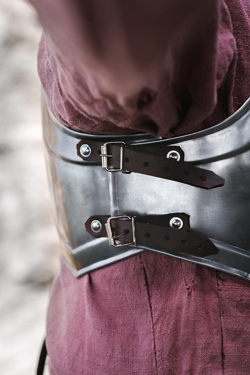 May include: Close-up of a silver metal chest plate with two brown leather straps and silver buckles. The chest plate is worn over a burgundy shirt.