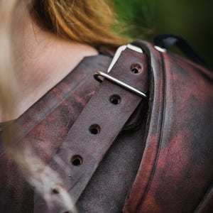 May include: Close-up of a brown leather shoulder armor piece with a silver buckle and strap. The leather has a distressed look and is worn.