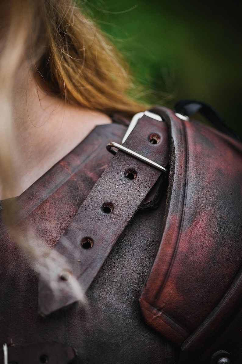 May include: Close-up of a brown leather shoulder armor piece with a silver buckle and strap. The leather has a distressed look and is worn.