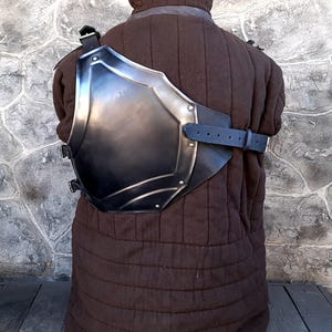 May include: A metal chest plate with a leather strap, worn over a brown quilted garment. The chest plate is curved and has a rounded shape.