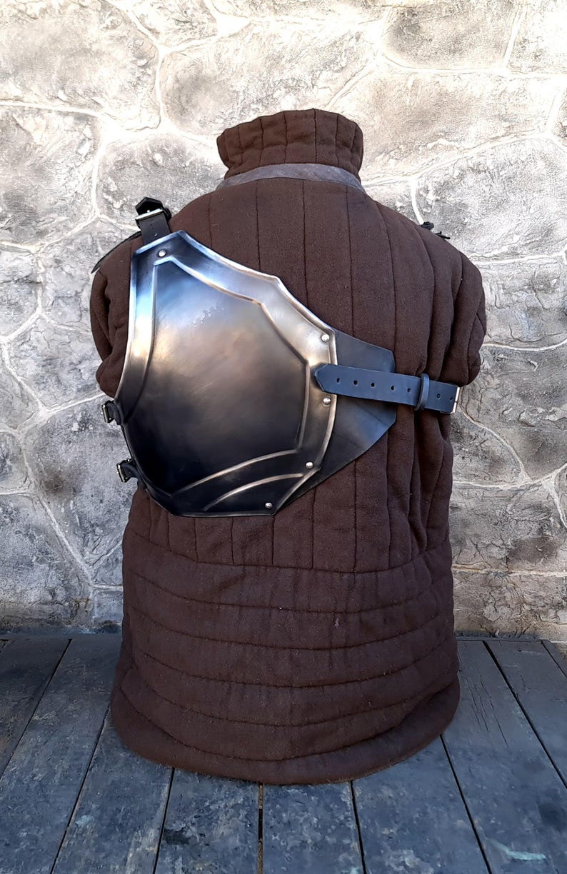 May include: A metal chest plate with a leather strap, worn over a brown quilted garment. The chest plate is curved and has a rounded shape.