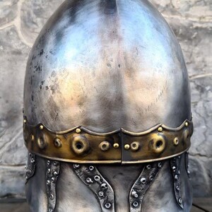 May include: A medieval-style helmet with a rounded top and a decorative brass band. The helmet is made of silver-colored metal and features riveted accents along the lower edge. The background is a stone wall.