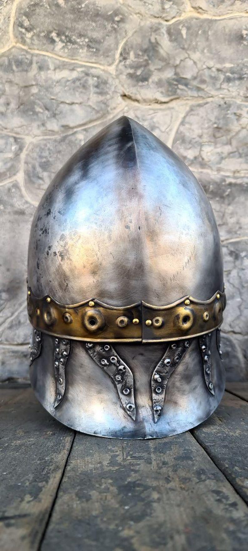 May include: A medieval-style helmet with a rounded top and a decorative brass band. The helmet is made of silver-colored metal and features riveted accents along the lower edge. The background is a stone wall.
