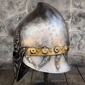 May include: A silver-toned medieval helmet with a rounded top and a gold-colored band around the base. The helmet has decorative rivets and a dark visor. The helmet is displayed on a black stand against a stone wall.