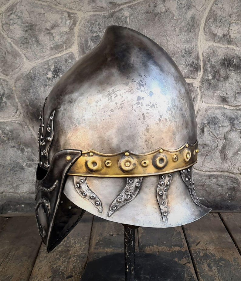 May include: A silver-toned medieval helmet with a rounded top and a gold-colored band around the base. The helmet has decorative rivets and a dark visor. The helmet is displayed on a black stand against a stone wall.