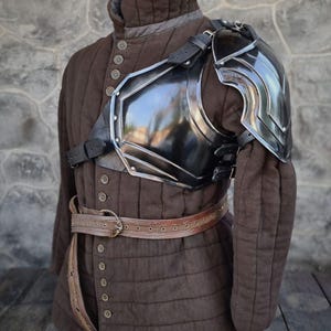 May include: A brown padded gambeson with a black metal shoulder pauldron and leather straps. The gambeson has a high collar and is buttoned down the front. A brown leather belt is also visible.