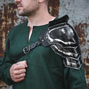 May include: A black metal shoulder armor piece with a leather strap. The armor is worn on the left shoulder and has a textured surface.