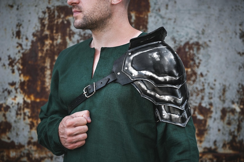 May include: A black metal shoulder armor piece with a leather strap. The armor is worn on the left shoulder and has a textured surface.