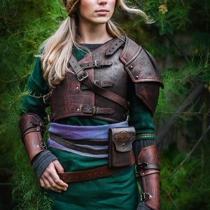 May include: A woman wearing a green tunic and brown leather armor with a belt and a pouch. She has long blonde hair and pointed ears.