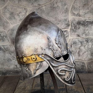 May include: A silver-colored medieval helmet with a pointed top and ornate detailing. The helmet features a gold-colored band around the base and decorative elements on the sides and front. The helmet is displayed on a black stand.