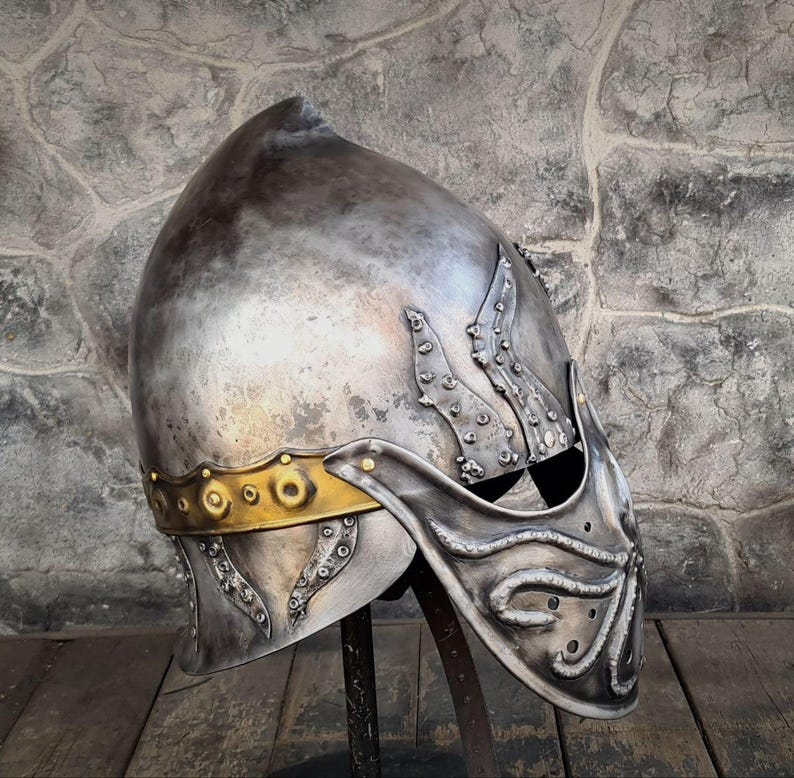 May include: A silver-colored medieval helmet with a pointed top and ornate detailing. The helmet features a gold-colored band around the base and decorative elements on the sides and front. The helmet is displayed on a black stand.