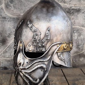 May include: A silver-colored metal helmet with an octopus design on the face guard and side. The helmet has a pointed top and a gold-colored band around the base. The octopus design features tentacles and suction cups.